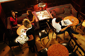 The picture shows a viennese coffeehouse seen from above. A waitress is carrying a tablet of coffee and water to a table where two women, seated in a velvet sofa, are talking. A man seated at the marble table next to them is reading a newspaper.