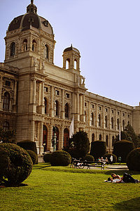 The picture shows people relaxing on the lawn in fron of the main entrance of the neo-baroque Museum of Art History