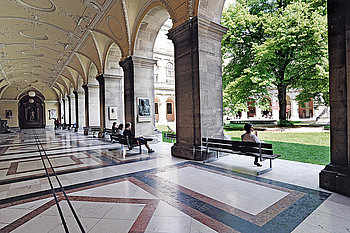 The picture shows the students sitting on benches under the arcades of the green courtyard of the main university building